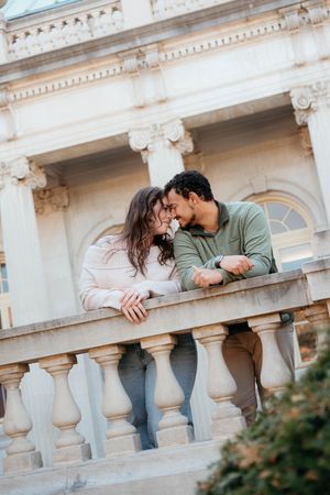 A couple shares a romantic moment while leaning over an ornate white stone balustrade of a classical building.