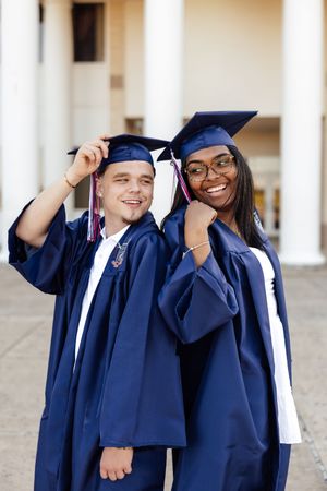Two graduates in blue caps and gowns smile proudly outside a university building on graduation day.