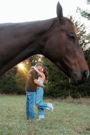 A couple embraces in a grassy field at sunset while a brown horse watches curiously in the background.