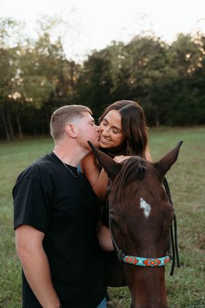 A couple shares a tender moment with their brown horse with a white star marking in a grassy field at sunset.