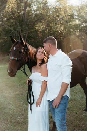A couple in a white dress and casual attire pose together with their brown horse in a sunlit field.