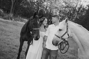 A couple in casual attire stands between two horses, one black and one white, in a grassy field.