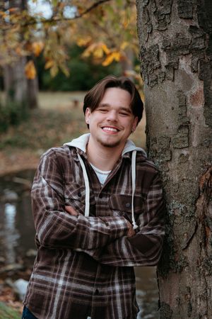 Person in plaid flannel shirt smiles while leaning against tree trunk during autumn season.