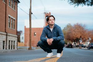 A person in a blue jacket sits thoughtfully on steps outside brick buildings on an autumn day.