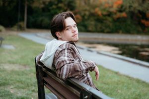 Young man sitting relaxed on a park bench outdoors, wearing a plaid shirt, with autumn trees in the background.