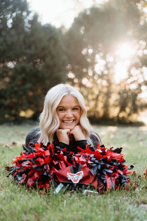 Cheerleader poses playfully in grass with red and black pom poms during golden hour sunset photoshoot.