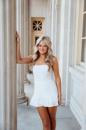 Woman in white mini dress poses elegantly in front of classical columns in outdoor architectural setting.