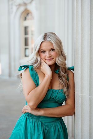 Woman in emerald green dress with ruffled straps poses against white architectural column with soft natural lighting.