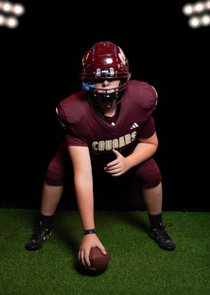 Young football player in maroon Cougars uniform crouches over a football under stadium lights on artificial turf.