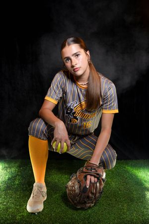 Young softball player in gray and gold uniform kneeling on grass, holding yellow ball and wearing catcher's mitt.