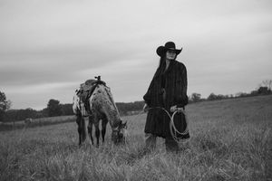 Black and white photograph of a figure in western attire with a horse grazing in a rural field under cloudy skies.