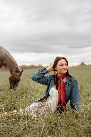 A person in a denim jacket and red scarf sits in a grassy field while a horse grazes in the background.
