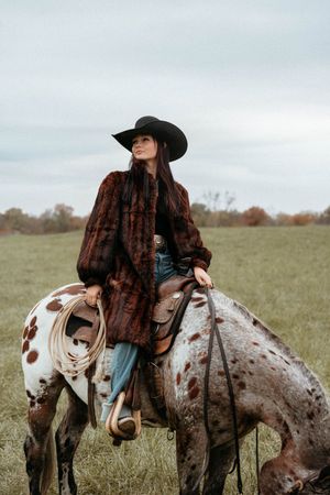 A spotted Appaloosa horse with brown and white markings stands in a grassy field with a rider in a black cowboy hat.