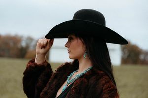 Woman in black hat and brown jacket poses in autumn field with dramatic lighting and moody atmosphere.