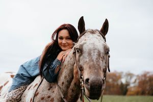 A person leans in close to a white horse's face while standing outdoors against an autumn background.