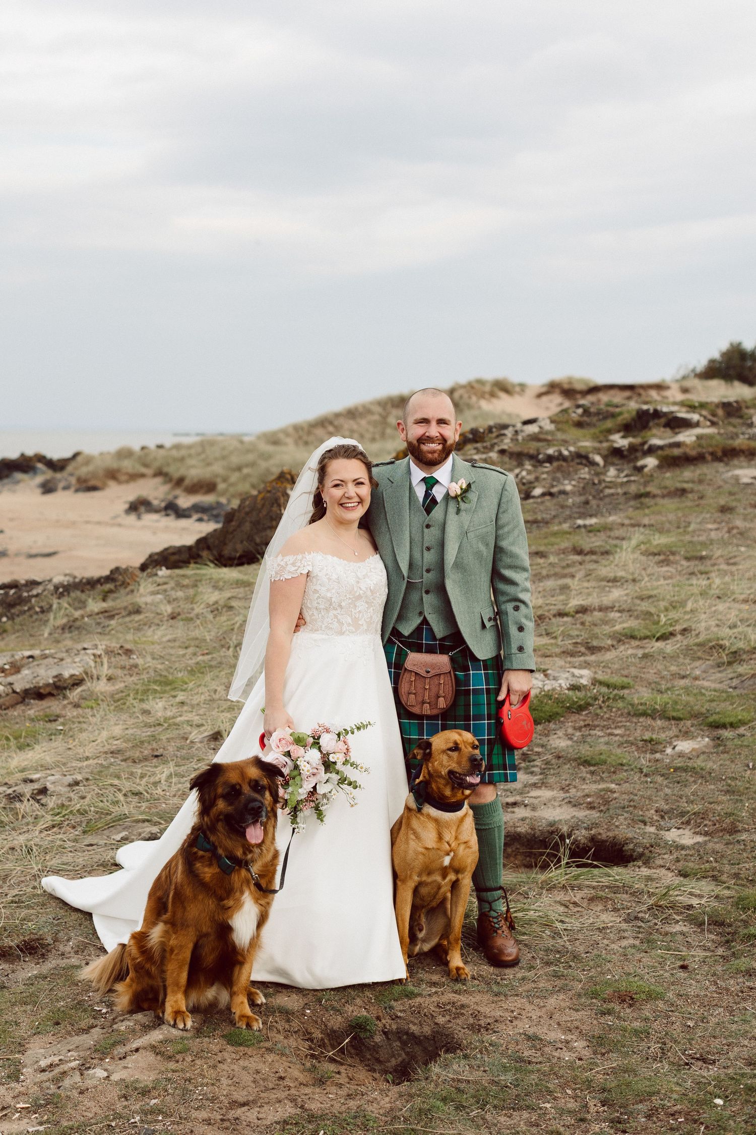 A couple in wedding attire poses on a hillside with two brown dogs in traditional Scottish highland dress.