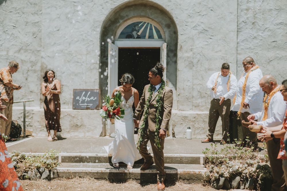 Military service member and person in white dress exit stone church building during sunny outdoor ceremony.