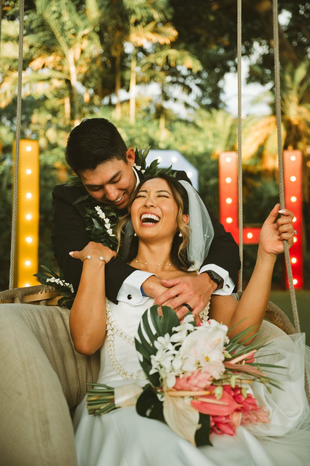 A joyful wedding couple laughs together while holding a bouquet of flowers in an outdoor garden setting.