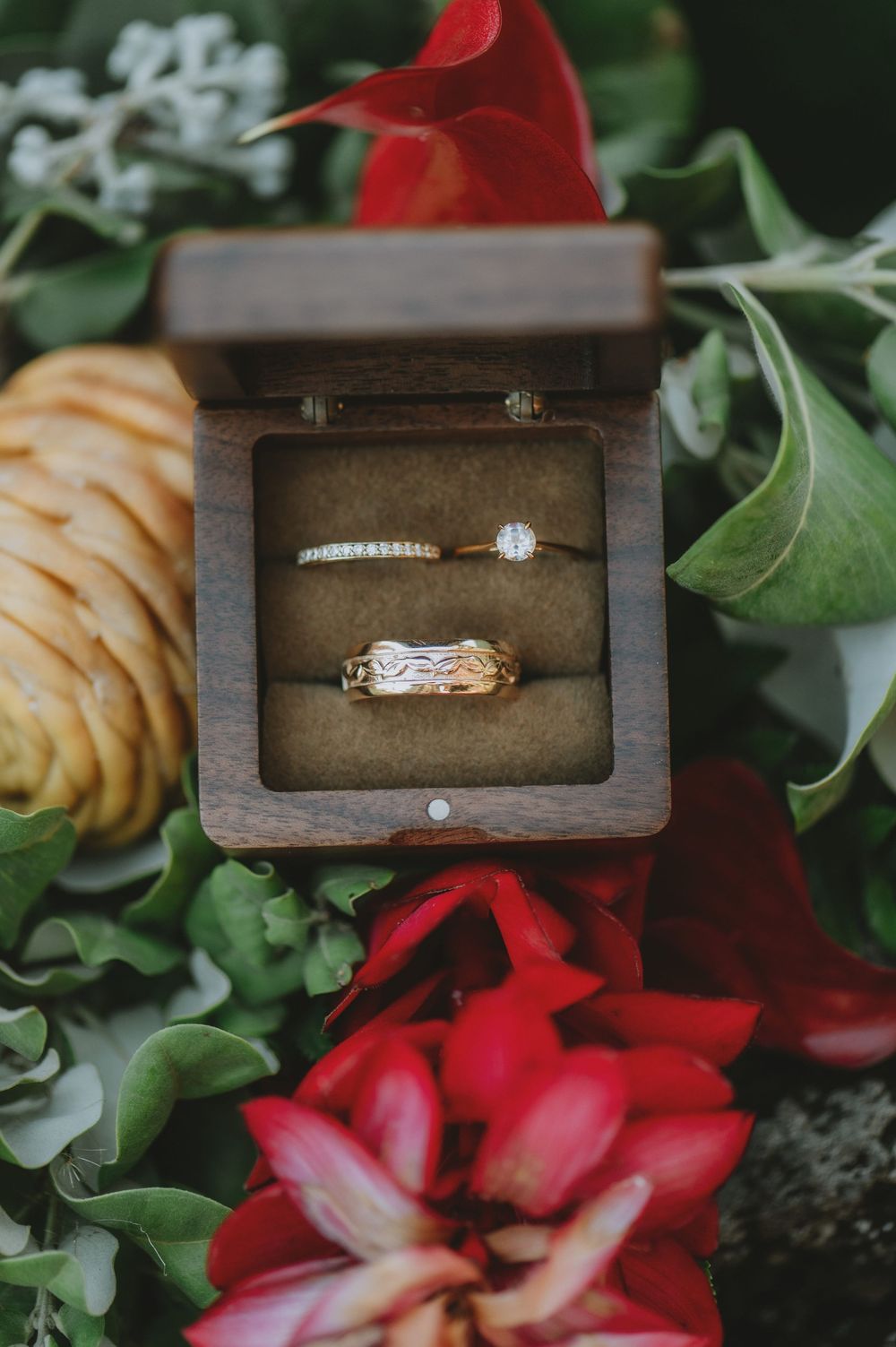 Diamond engagement ring displayed in wooden box with red flowers and greenery in romantic wedding detail shot.