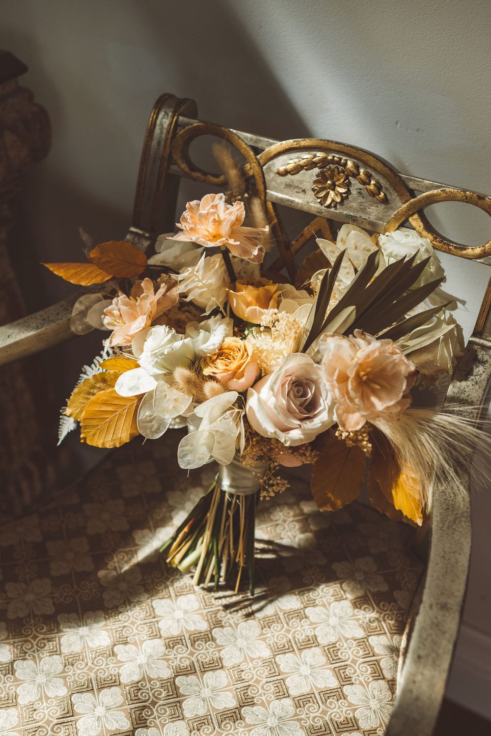 A romantic floral bouquet with peach roses and gold accents arranged on a textured lace tablecloth in soft lighting.