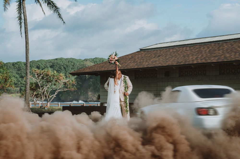 A bride in a white wedding dress walks along a dirt road with palm trees and mountains in the background.