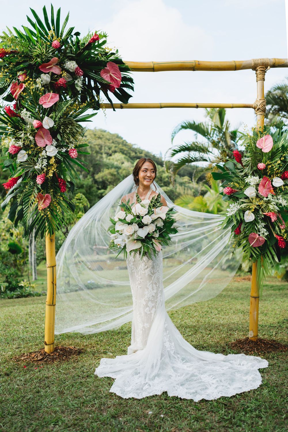 A tropical wedding arch decorated with lush greenery, burgundy and pink flowers against a scenic mountain backdrop.