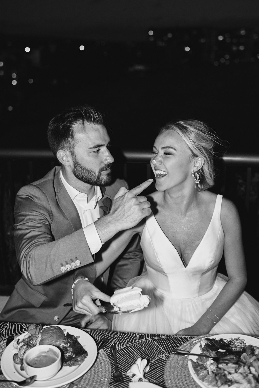 A couple in formal attire shares a joyful moment while dining together at a table in black and white photography.