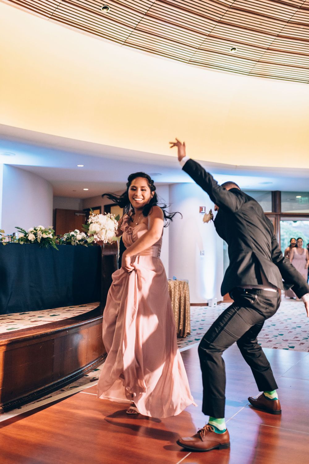Wedding party members perform a synchronized dance routine in a reception hall with hardwood floors and curved ceiling.