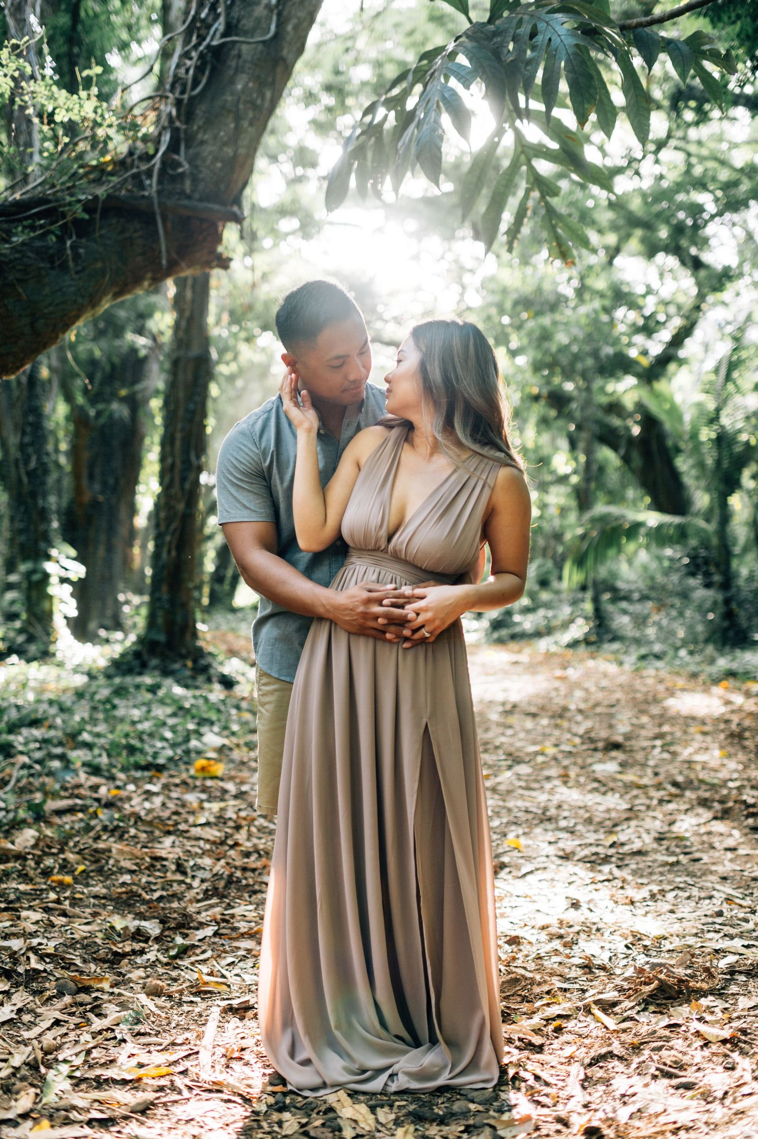 A couple embraces on a sunlit forest path surrounded by fallen leaves and trees.