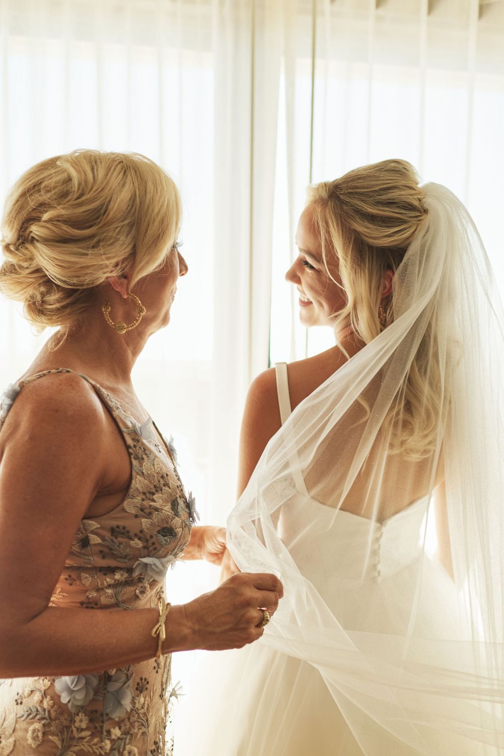 A tender moment as bridesmaids help adjust a flowing white wedding veil near a bright window.