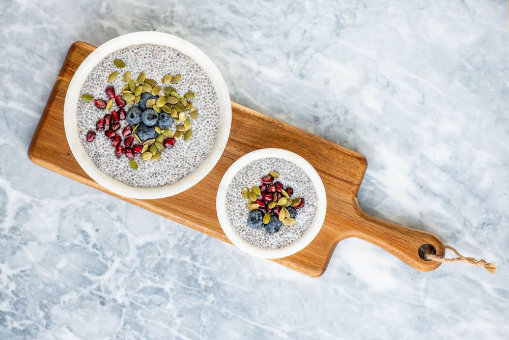 White chia pudding bowls topped with colorful seeds and berries on wooden serving board.