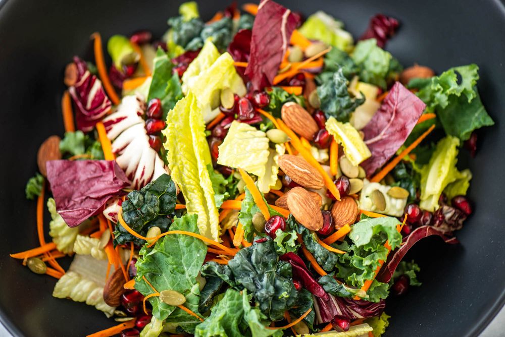 Vibrant kale salad with nuts, carrots and pomegranate seeds in black bowl.