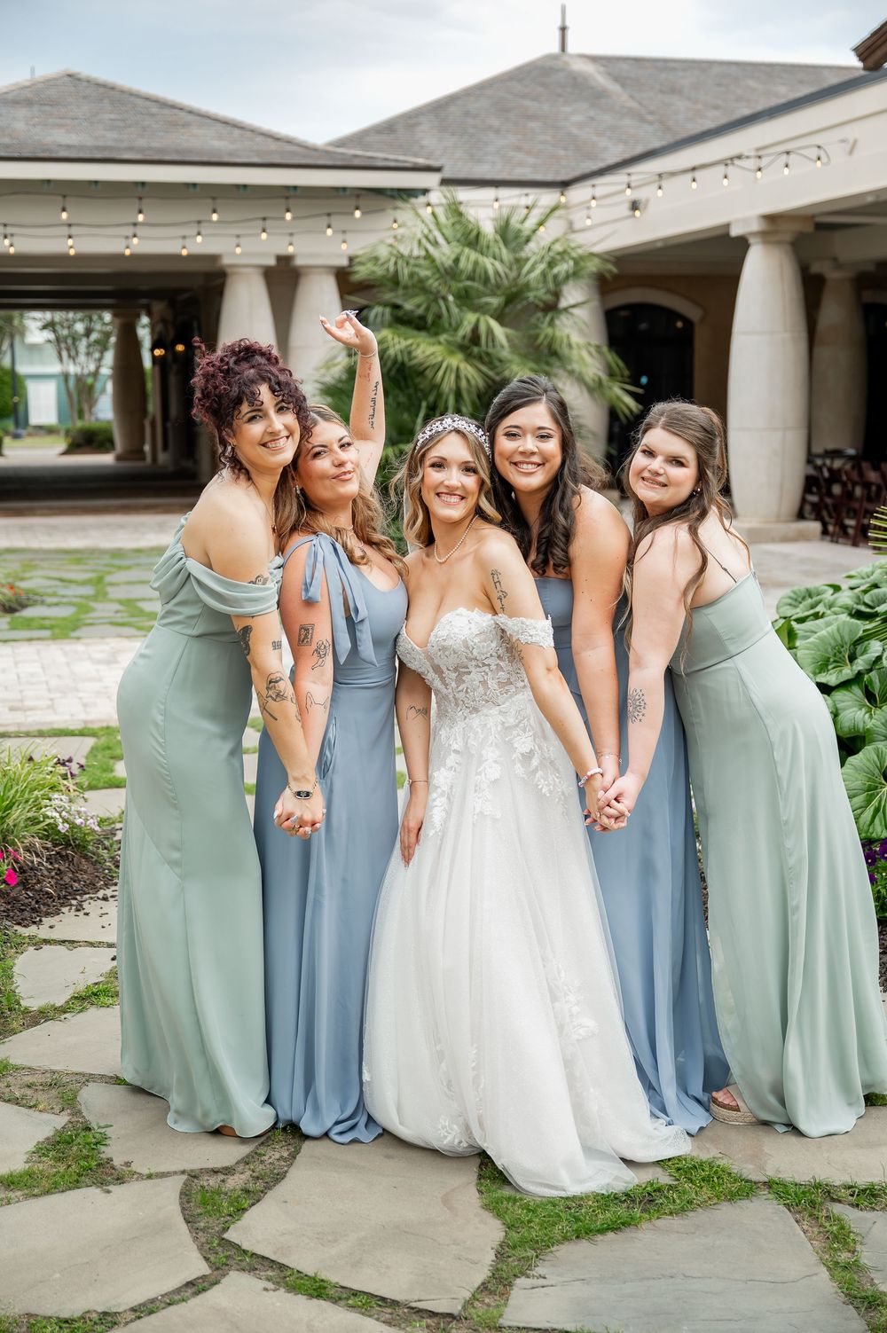 Wedding party in coordinated mint and blue dresses poses together on a covered patio.
