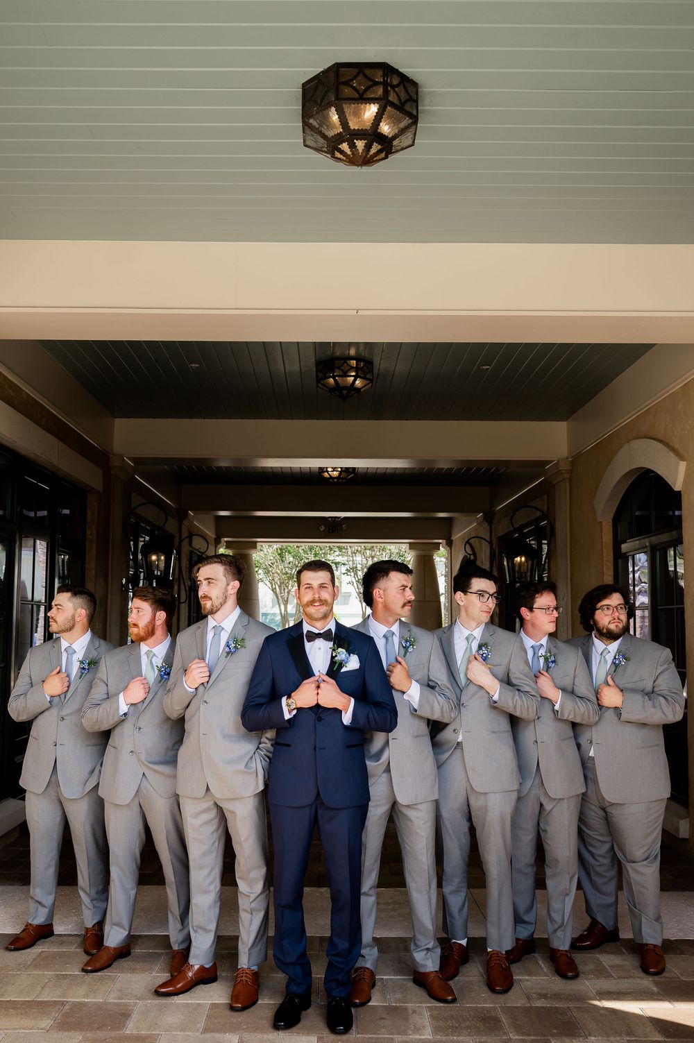 Groomsmen in light gray suits pose with groom in navy blue tuxedo under elegant ceiling.