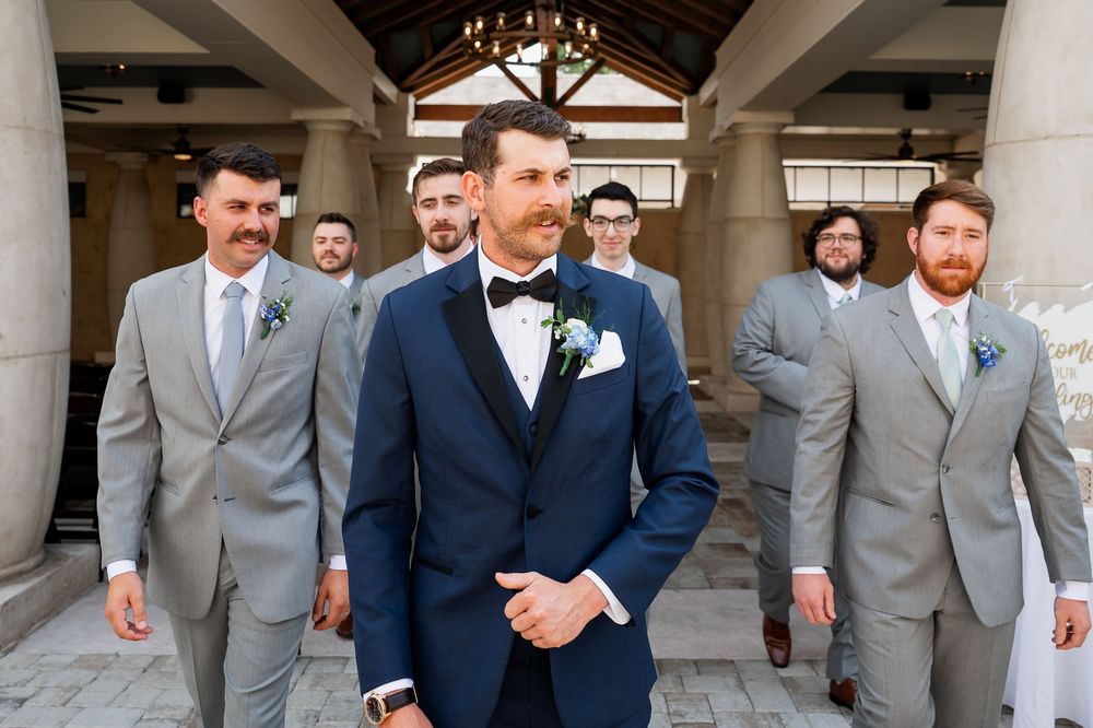 Wedding party members in suits gather for formal photos under a covered walkway.