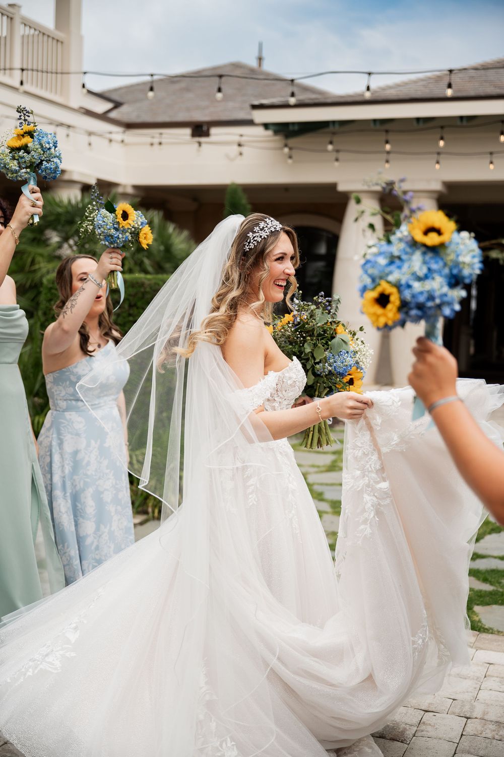 Bridesmaids in pastel dresses help arrange the bride's gown while holding sunflower bouquets.