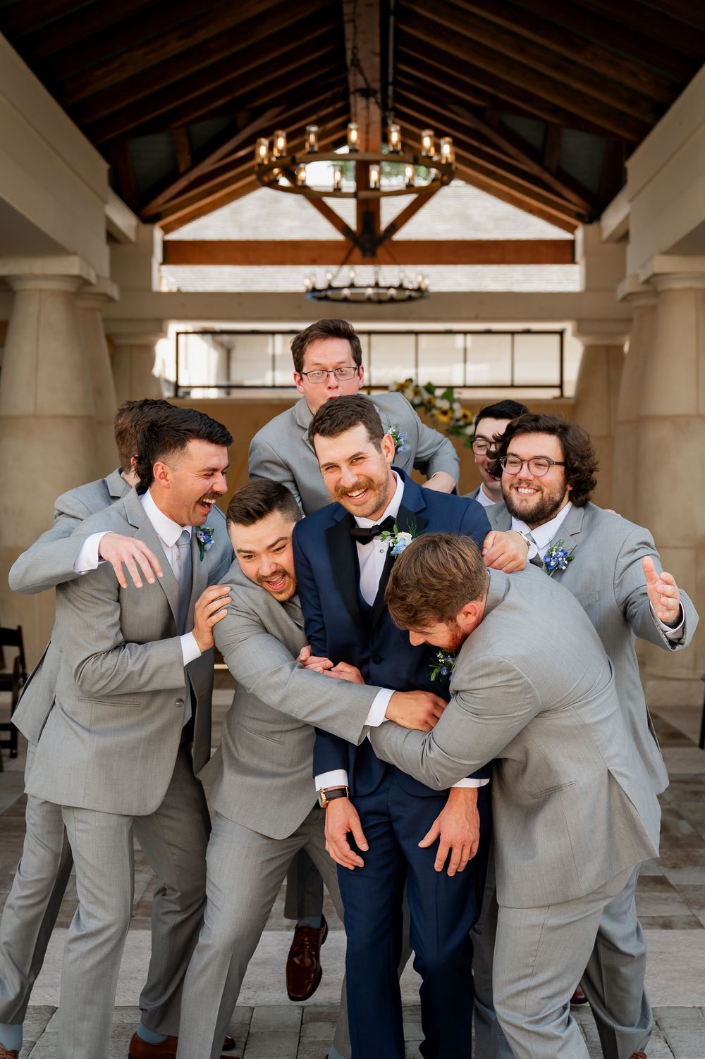 Groomsmen in gray suits playfully huddle around groom in navy tuxedo during wedding photos.