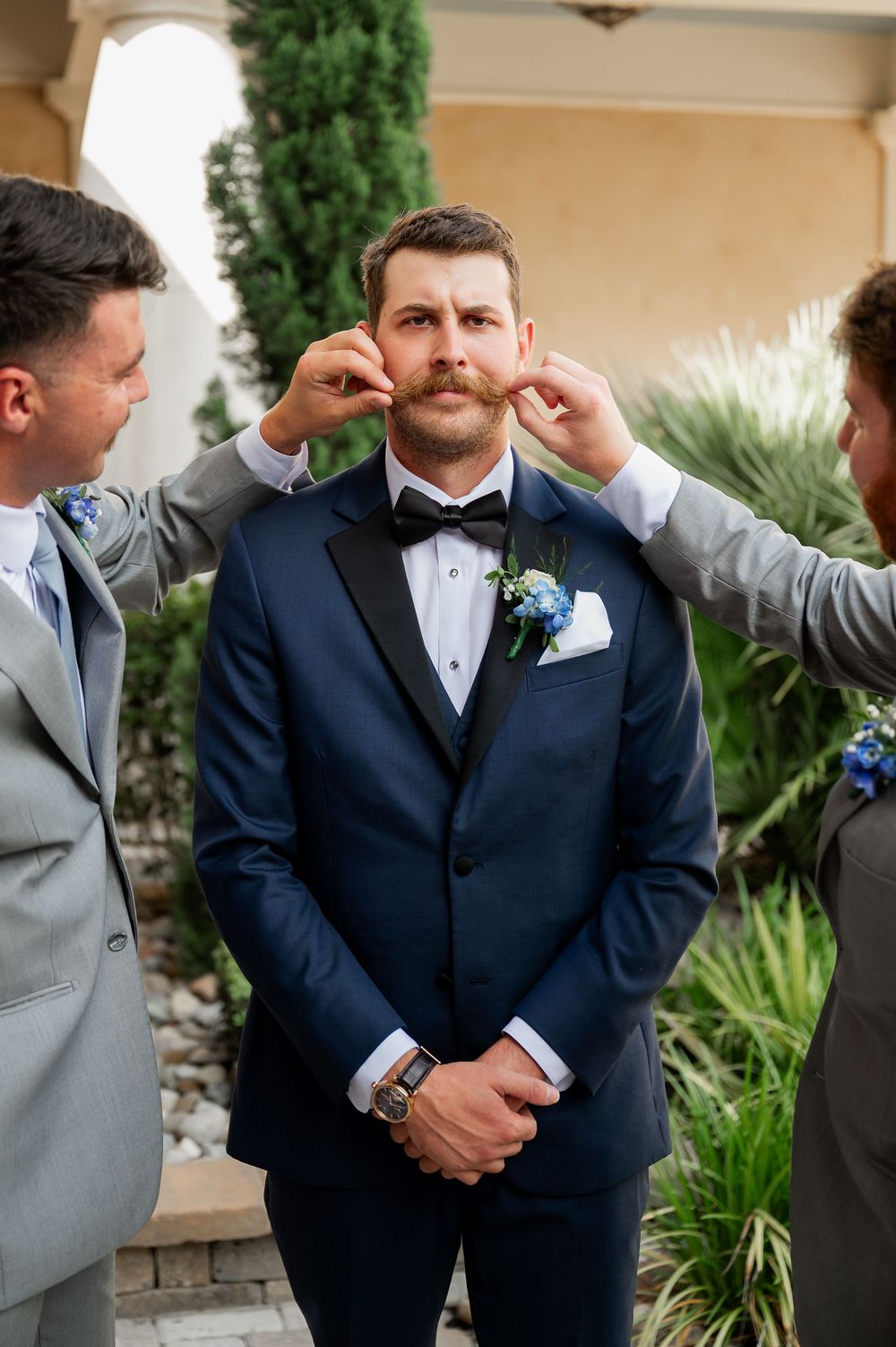 Groomsmen help adjust a navy blue tuxedo and bowtie during wedding preparations.