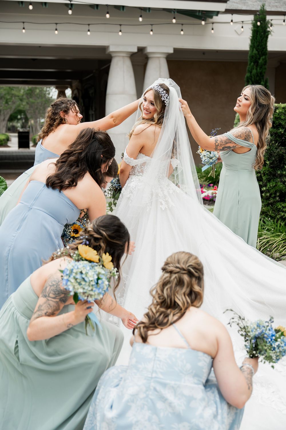 Bridesmaids in mint and blue dresses help adjust the bride's flowing white gown.