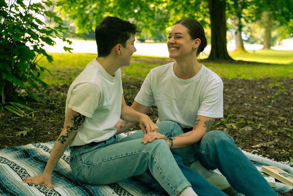 Two people in white t-shirts and jeans sit together outdoors on a sunny day surrounded by greenery.