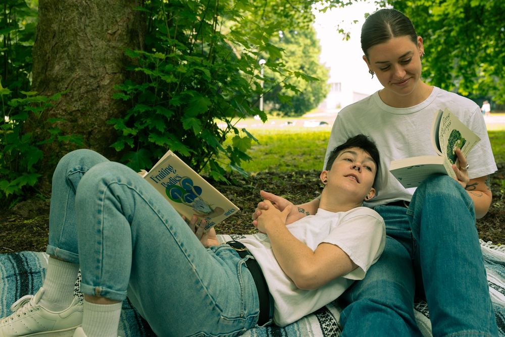 Students relax outdoors while reading books and studying under the shade of green trees on a sunny day.