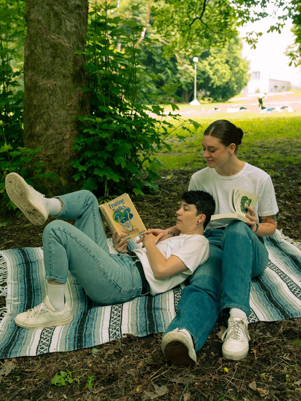 A group relaxes on a striped Mexican blanket under trees while reading books on a sunny afternoon.