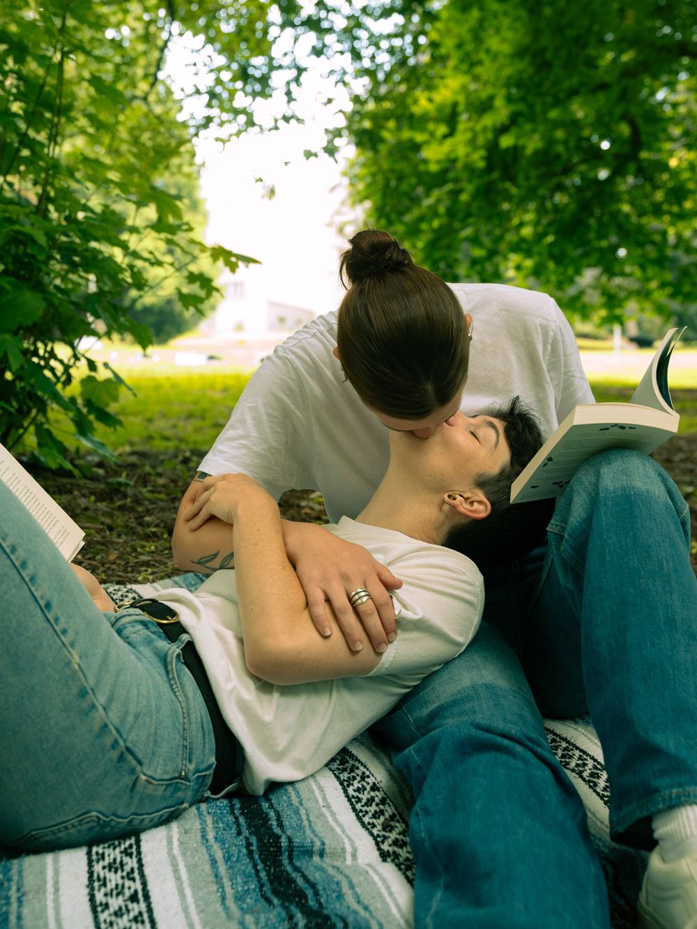 A couple relaxes together on a blanket in a park setting with lush green trees in the background.