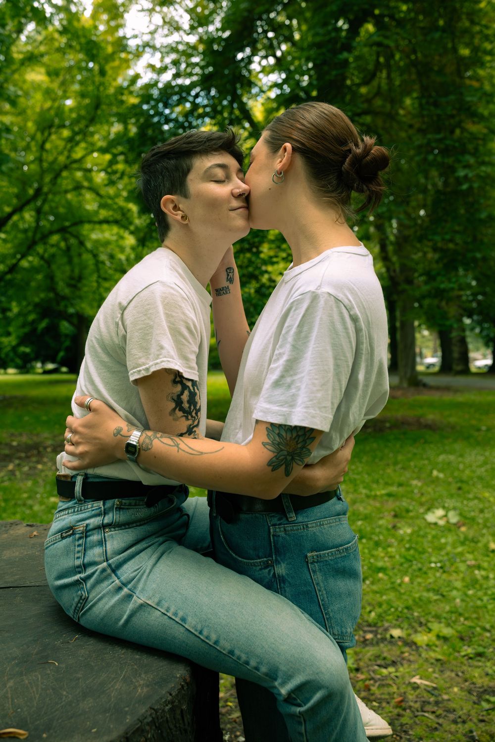 A couple in matching white t-shirts and jeans share intimate moments in a lush green park setting.