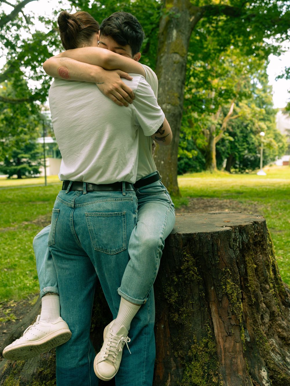 Two people in denim and white shirts share intimate moments near a tree stump in a park setting.
