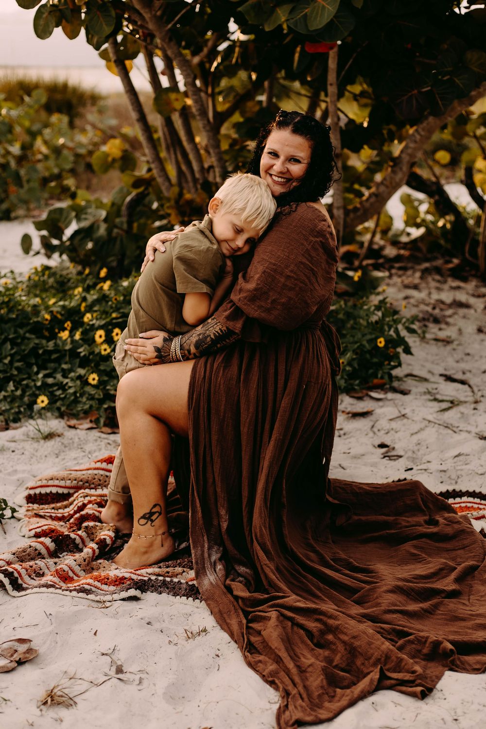 A romantic beach photoshoot featuring a couple wrapped in a flowing brown dress sitting on sandy shores.