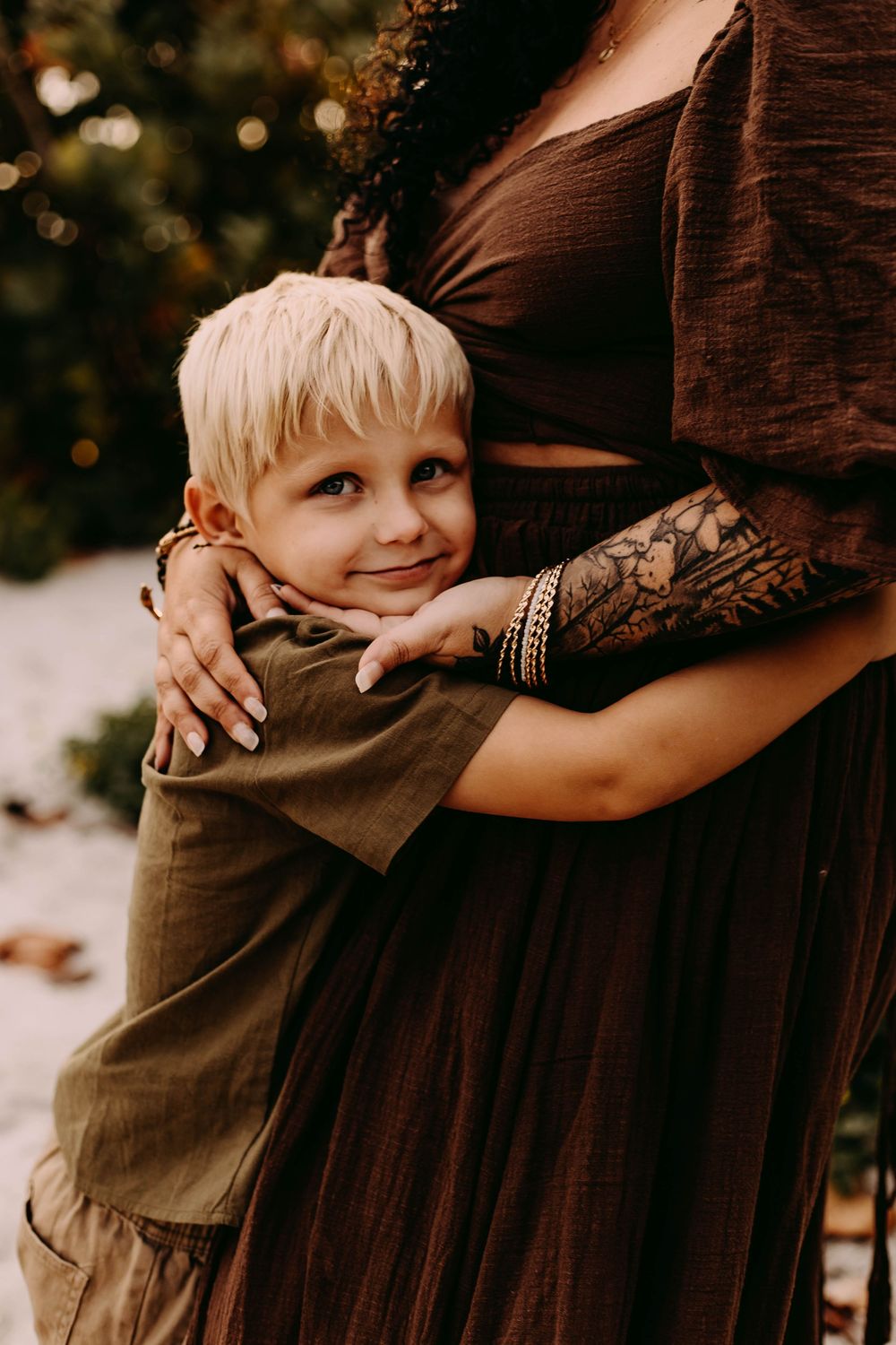 A child hugs a woman wearing a brown dress with lace sleeves in a warm autumn setting.