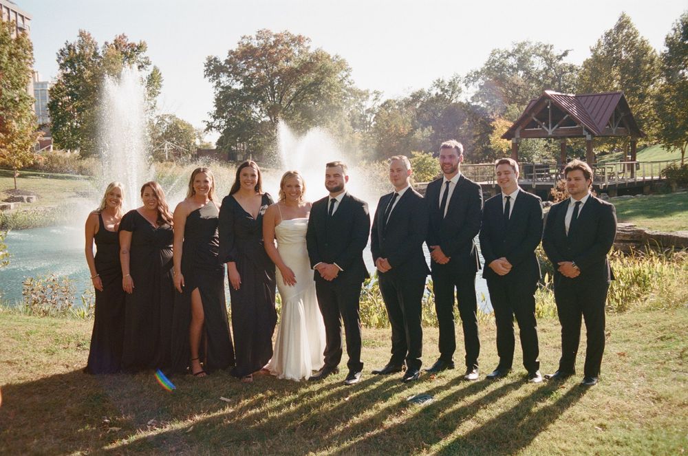 Wedding party poses outdoors in formal attire with sunlight creating silhouettes against a scenic natural backdrop.