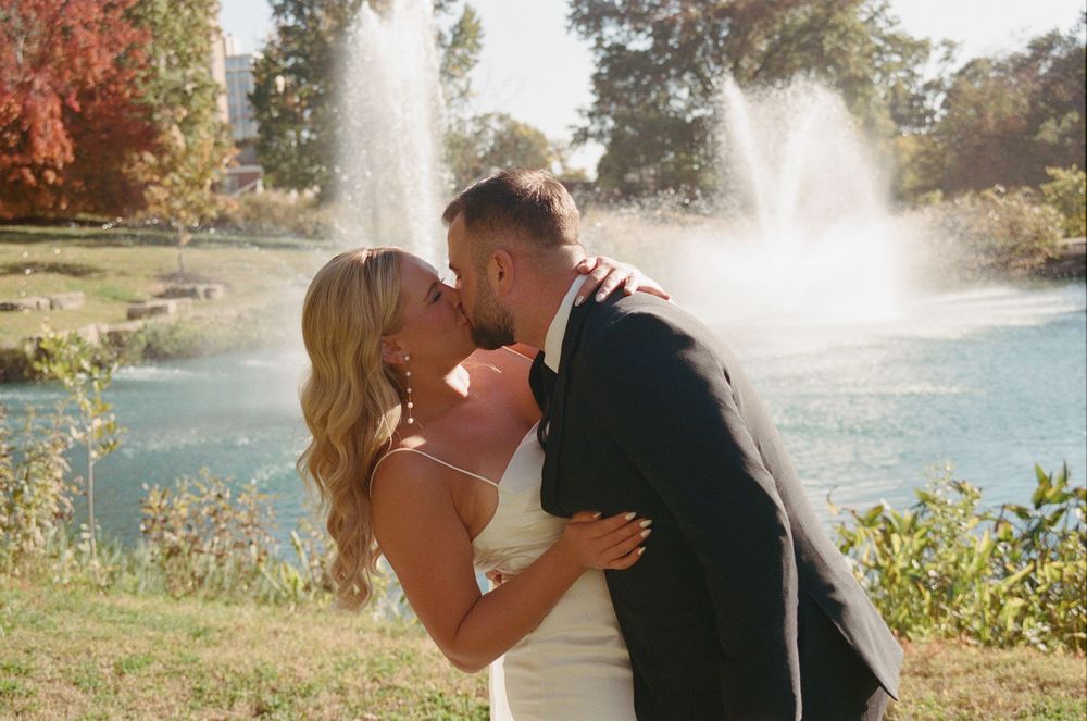 A romantic wedding kiss in front of a fountain with autumn foliage in the background.