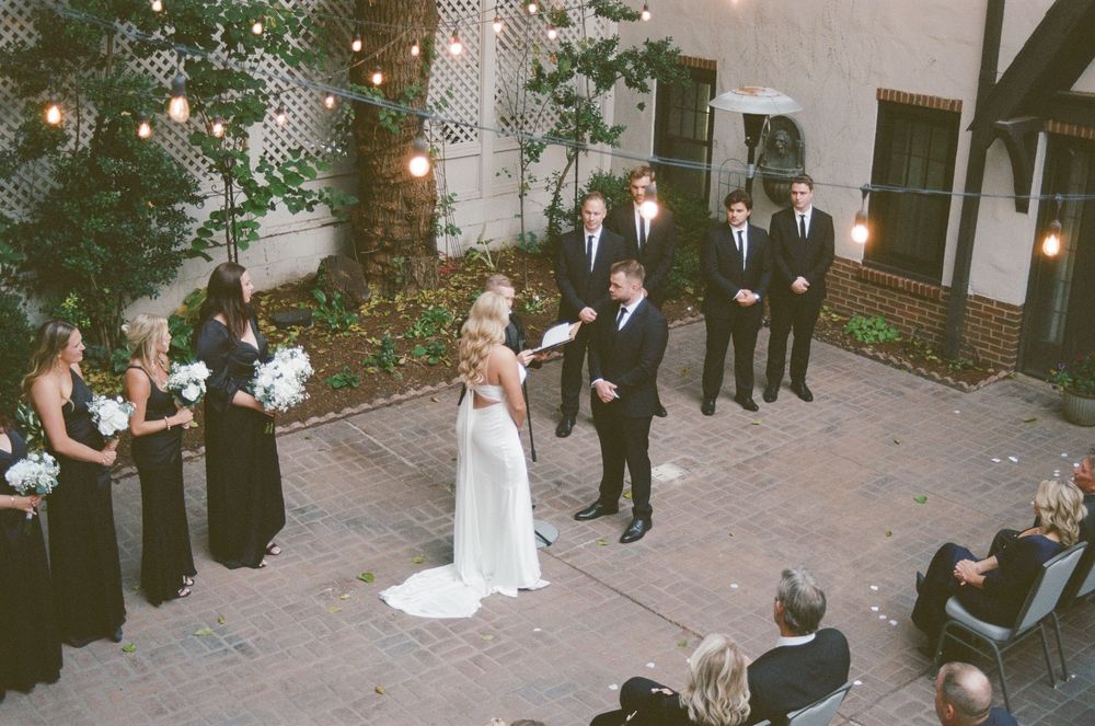 Wedding guests gather outside a church in a casual setting with string lights overhead and brick walls in the background.